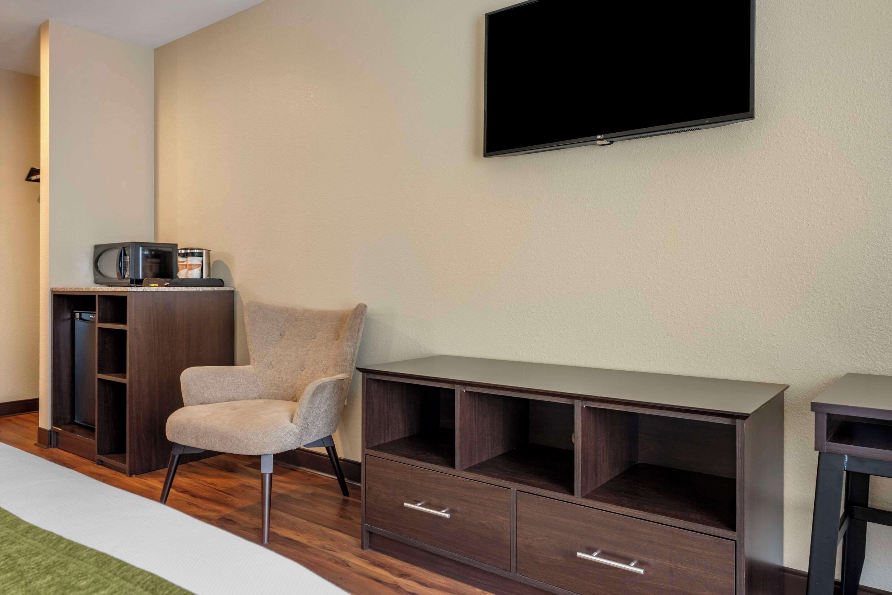 A hotel room with a flat-screen TV mounted on the wall, a wooden cabinet with drawers, a cushioned chair, and a small table.