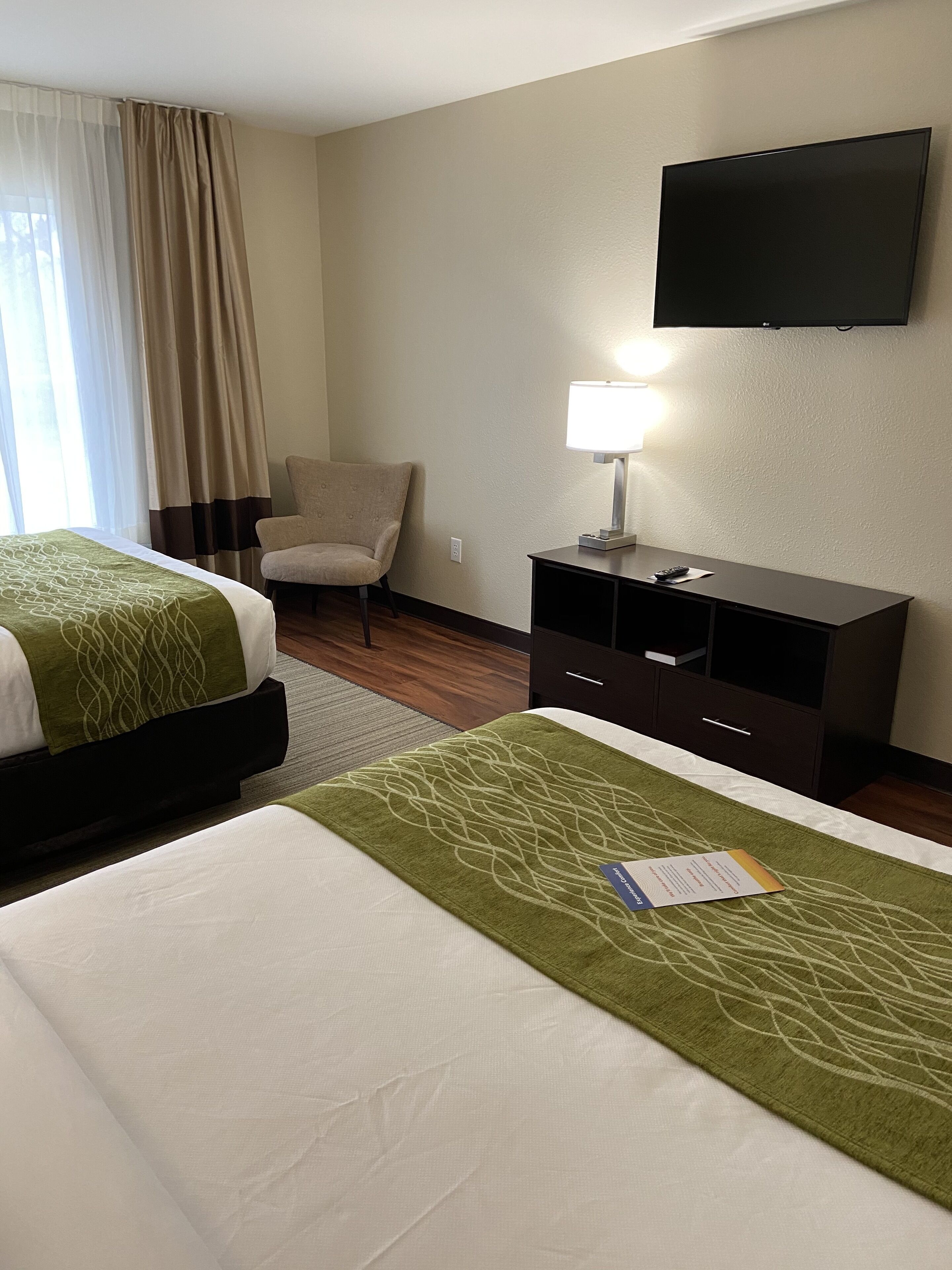 A hotel room with two beds, a chair, a desk with a lamp, and a TV on the wall.