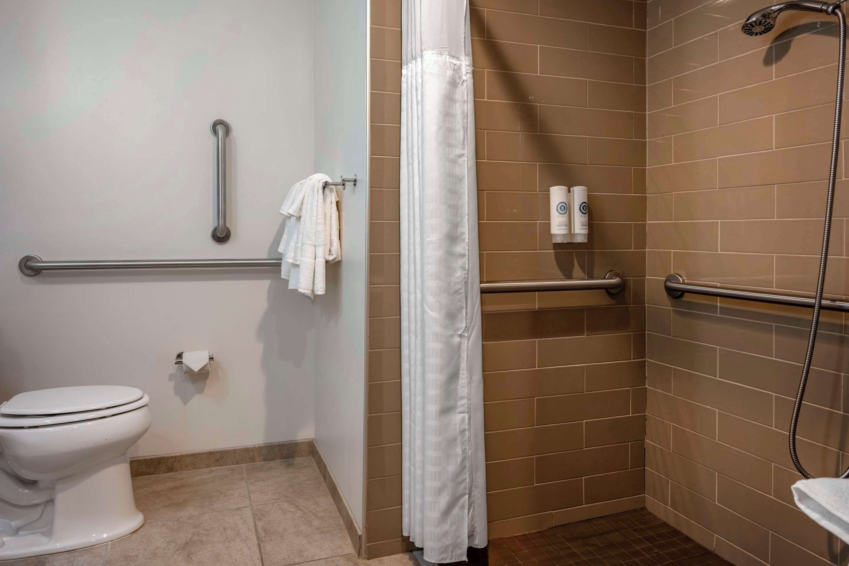 Baño con inodoro, ducha con cabezal de ducha de mano y barras de apoyo.