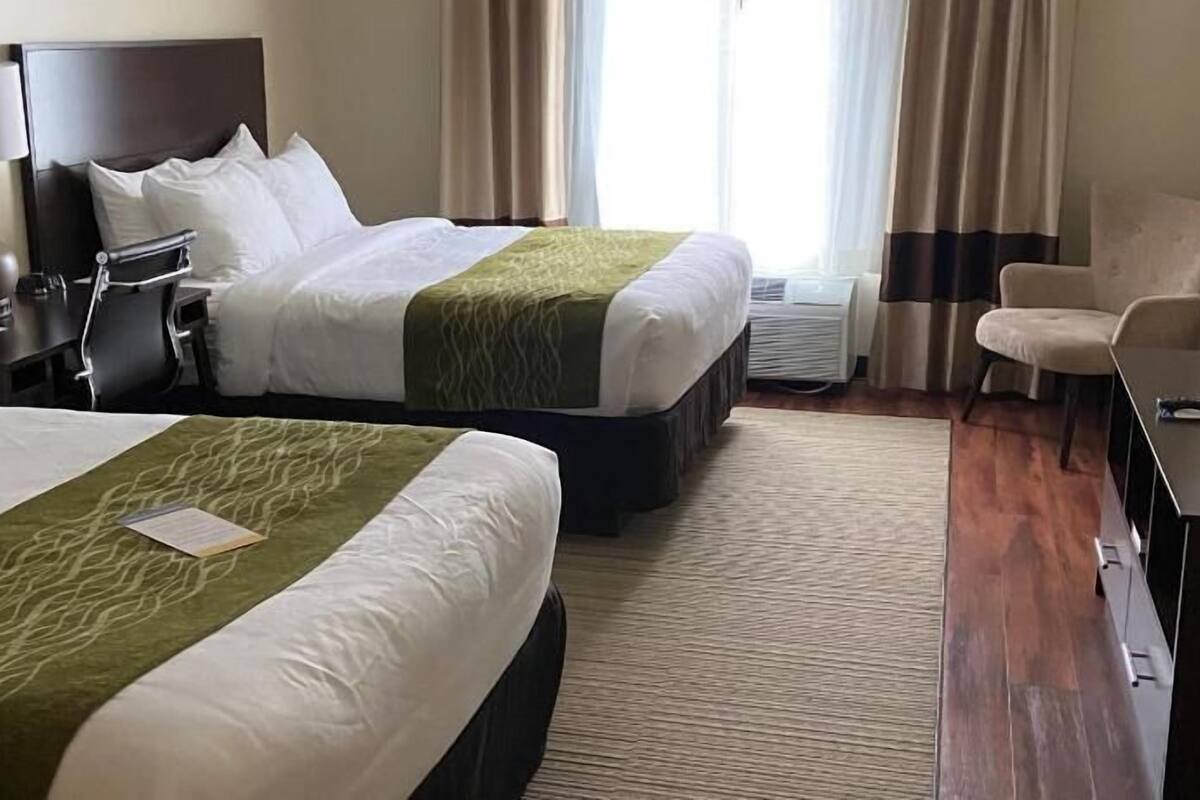 A hotel room with two beds, a desk, a chair, and a lamp.