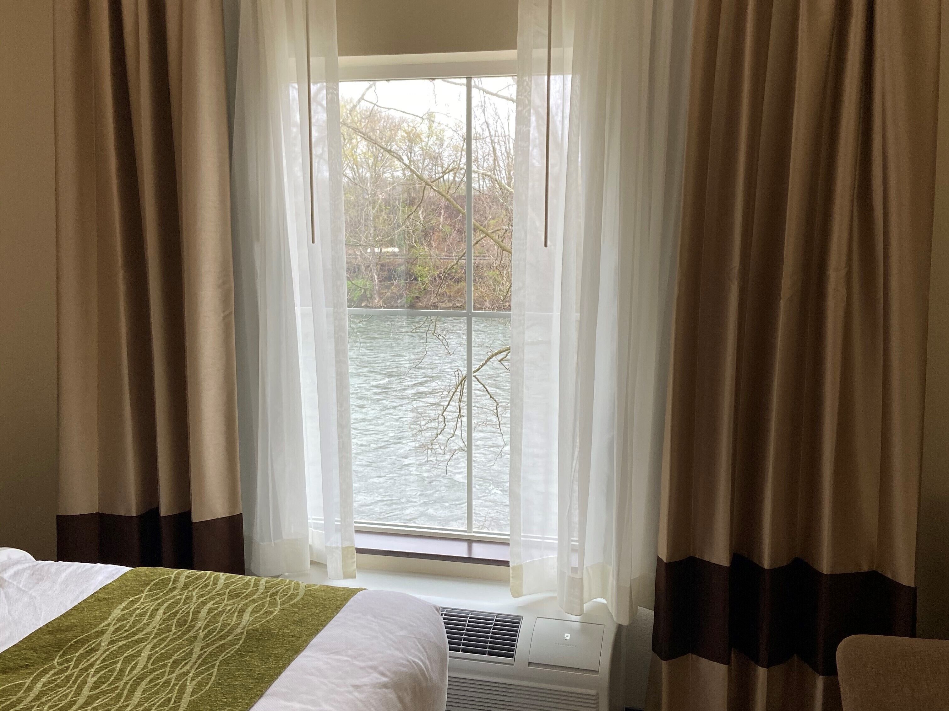 A hotel room with a bed, a window with curtains, and a view of a body of water and trees.