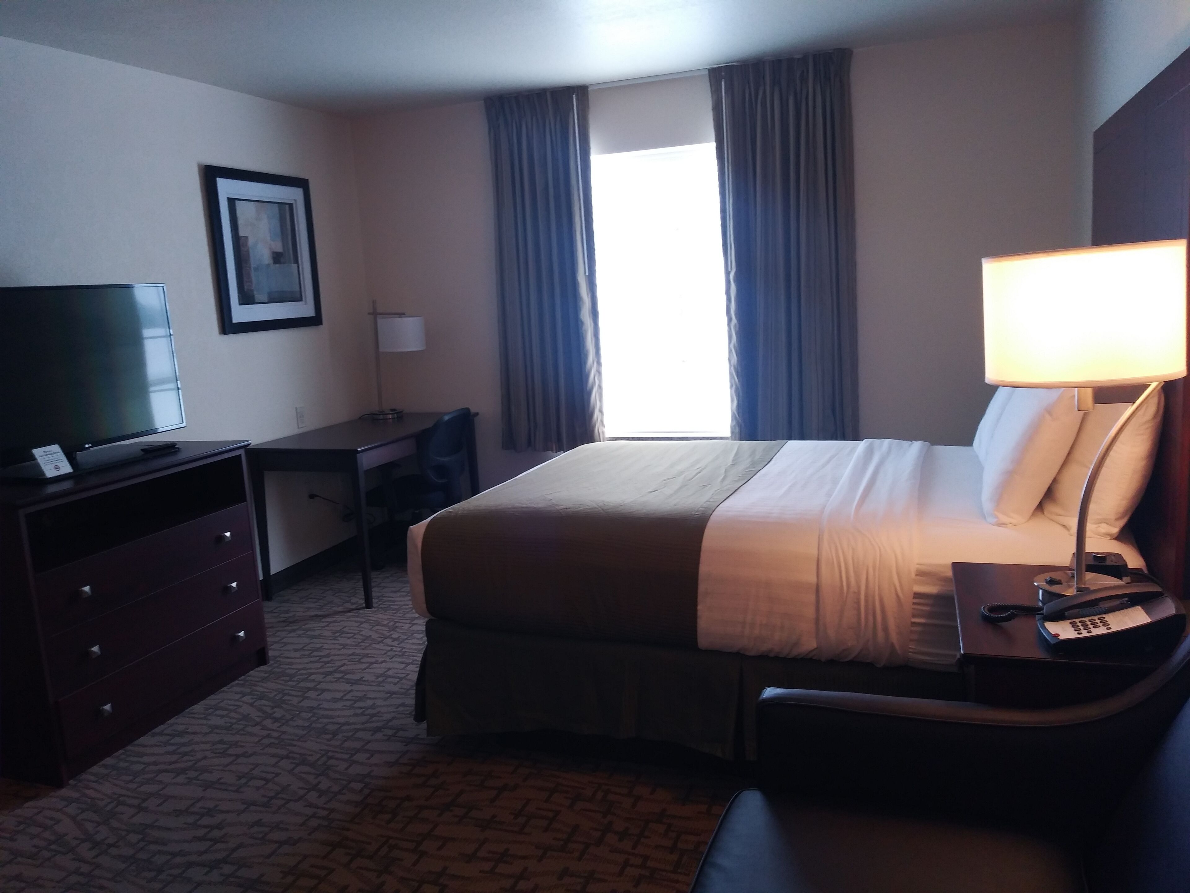 Quarto de hotel com cama, escrivaninha, cadeira, televisão, luminária e cortinas.