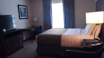 Quarto de hotel com cama, escrivaninha, cadeira, televisão, luminária e cortinas.