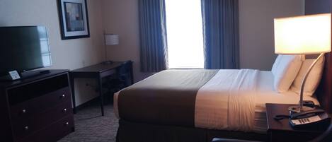 A hotel room with a bed, desk, chair, television, lamp, and curtains.