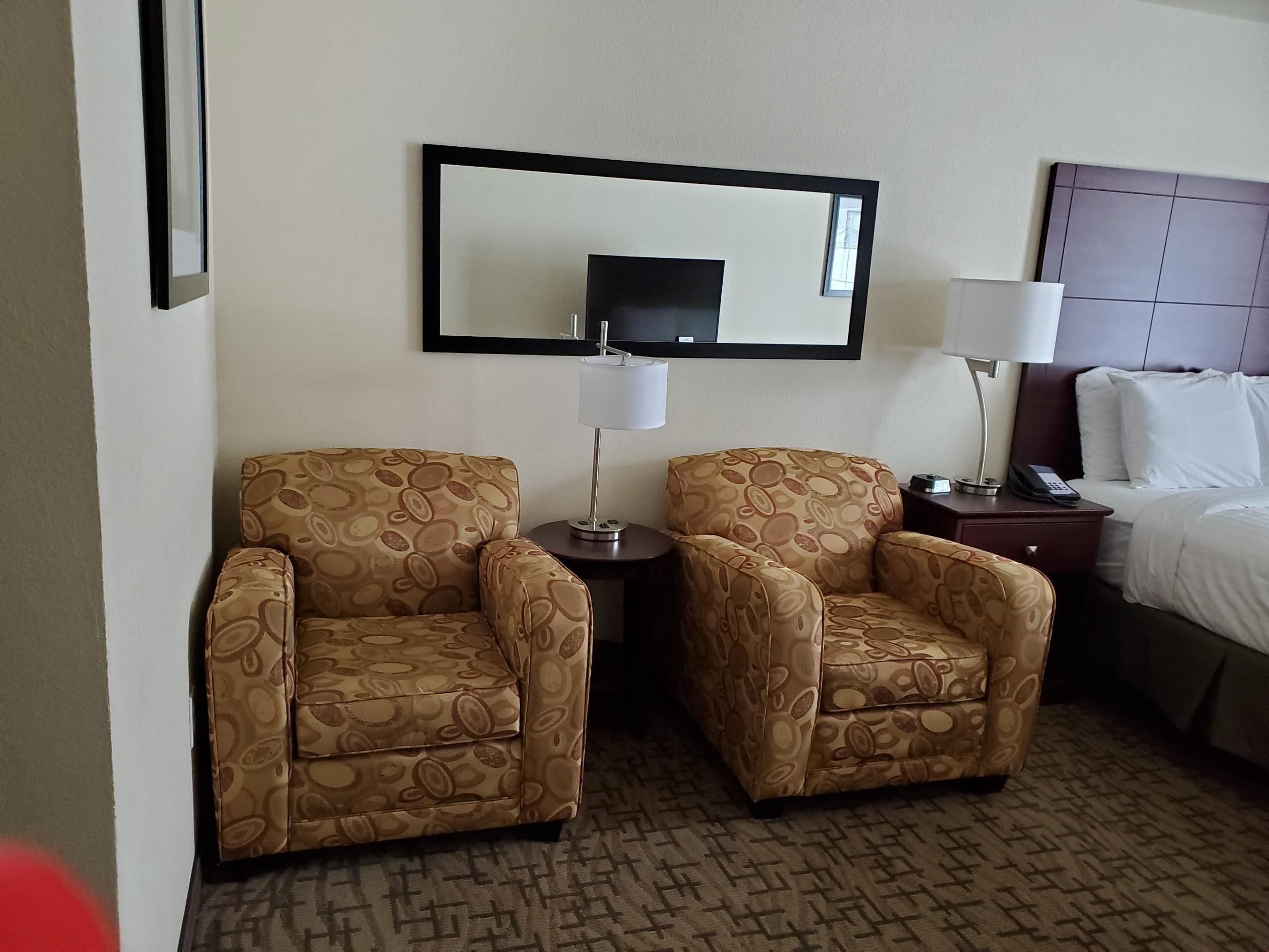 Habitación de hotel con dos sillones estampados, una mesita con lámpara, cama con ropa blanca y un espejo grande en la pared.
