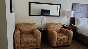 Habitación de hotel con dos sillones estampados, una mesita con lámpara, cama con ropa blanca y un espejo grande en la pared.