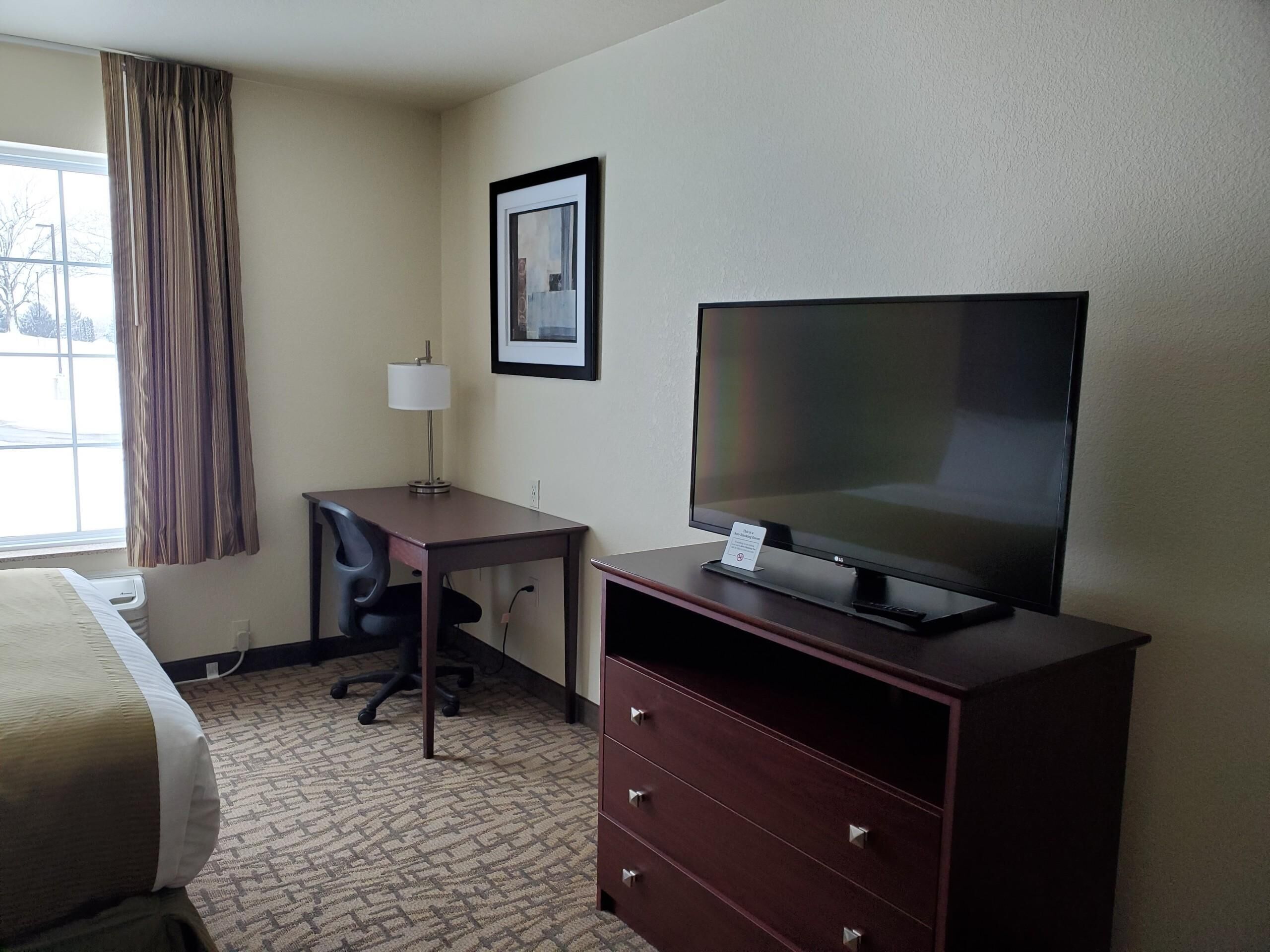 A hotel room with a bed, a desk with a chair, a television on a stand, a lamp, and a dresser.