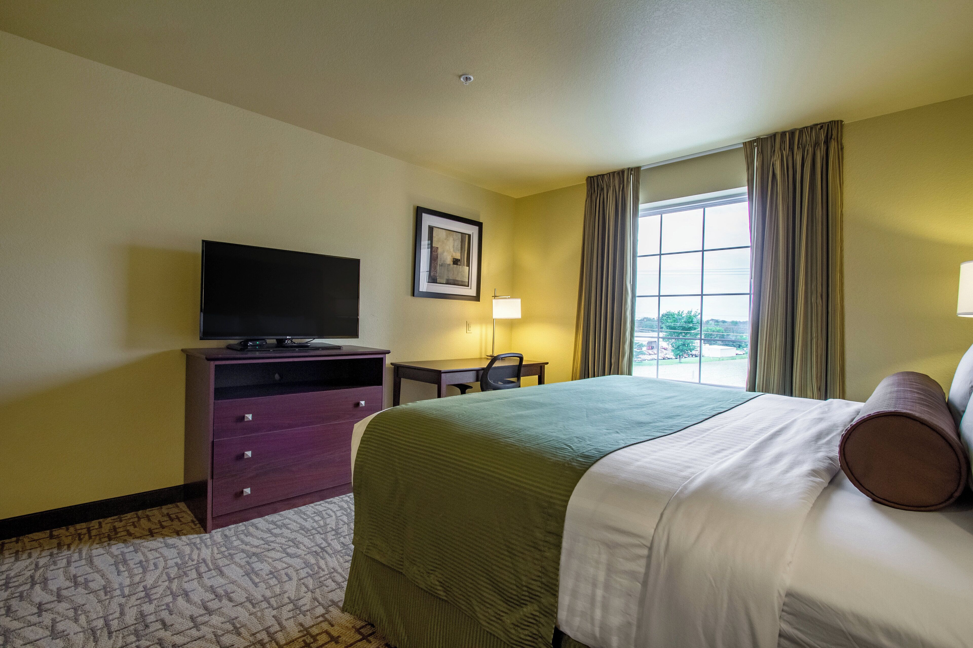 A hotel room with a bed, a television on a dresser, a desk with a chair, and a window with curtains.