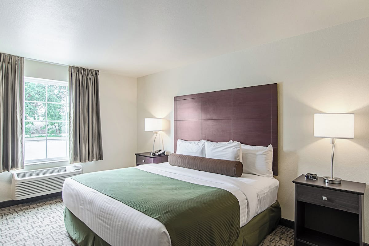 A hotel room with a large bed, two bedside tables, a window with curtains, a wall-mounted heater, and a nightstand with a lamp.