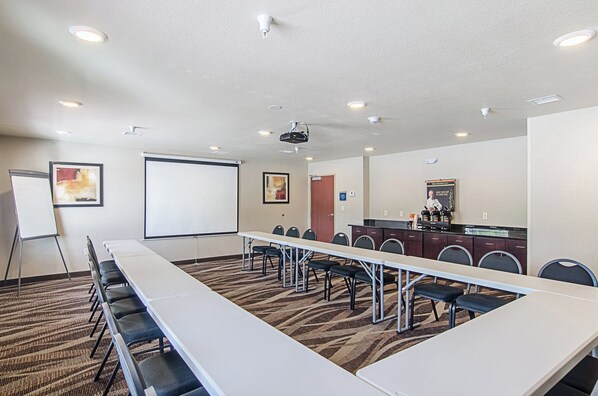 A conference room with a long table, chairs, a projector screen, and a small kitchenette.