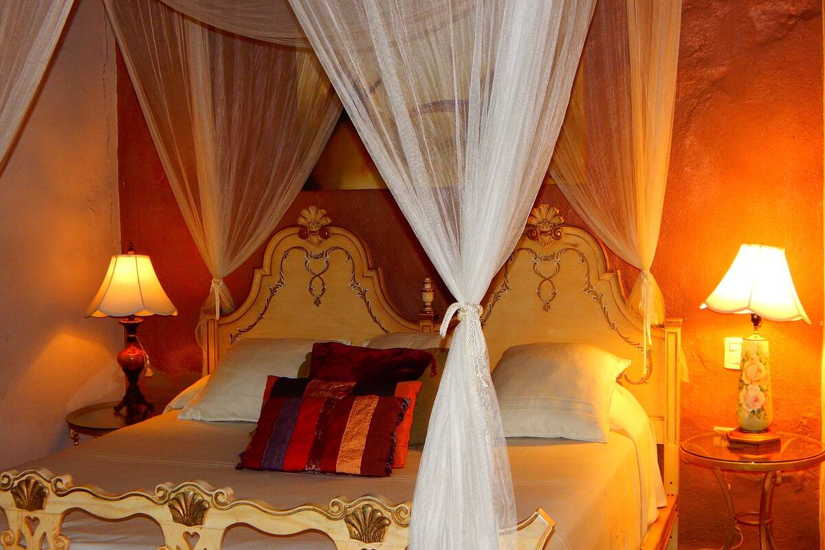 Een bed met hemel, twee bedlampjes en een nachtkastje met een lamp.
