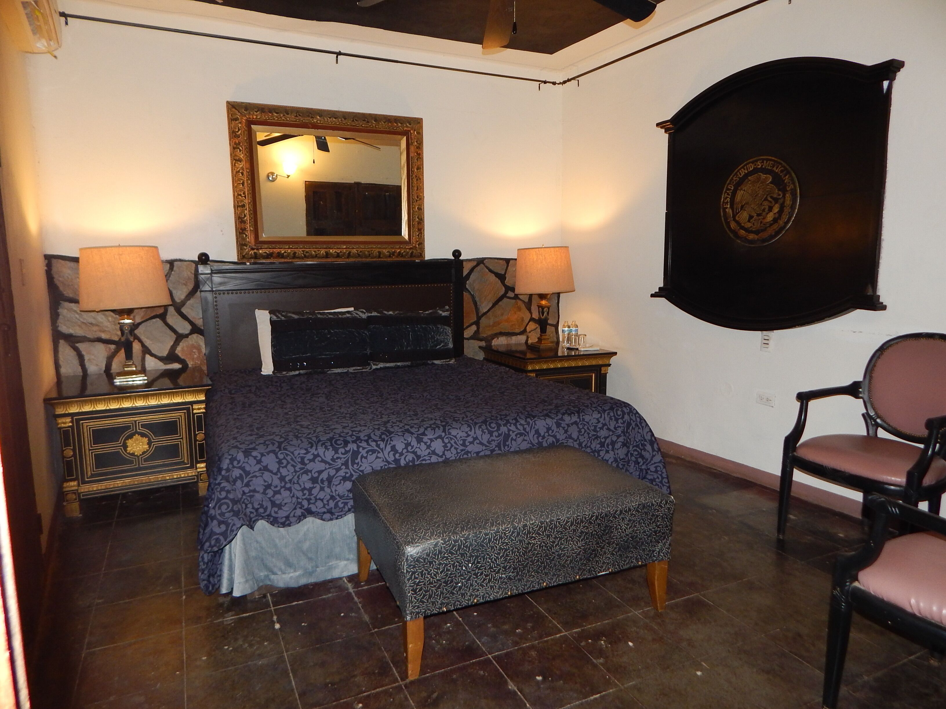 Un dormitorio con una cama, dos mesitas de noche con lámparas, un banco, una silla y un espejo grande en la pared.