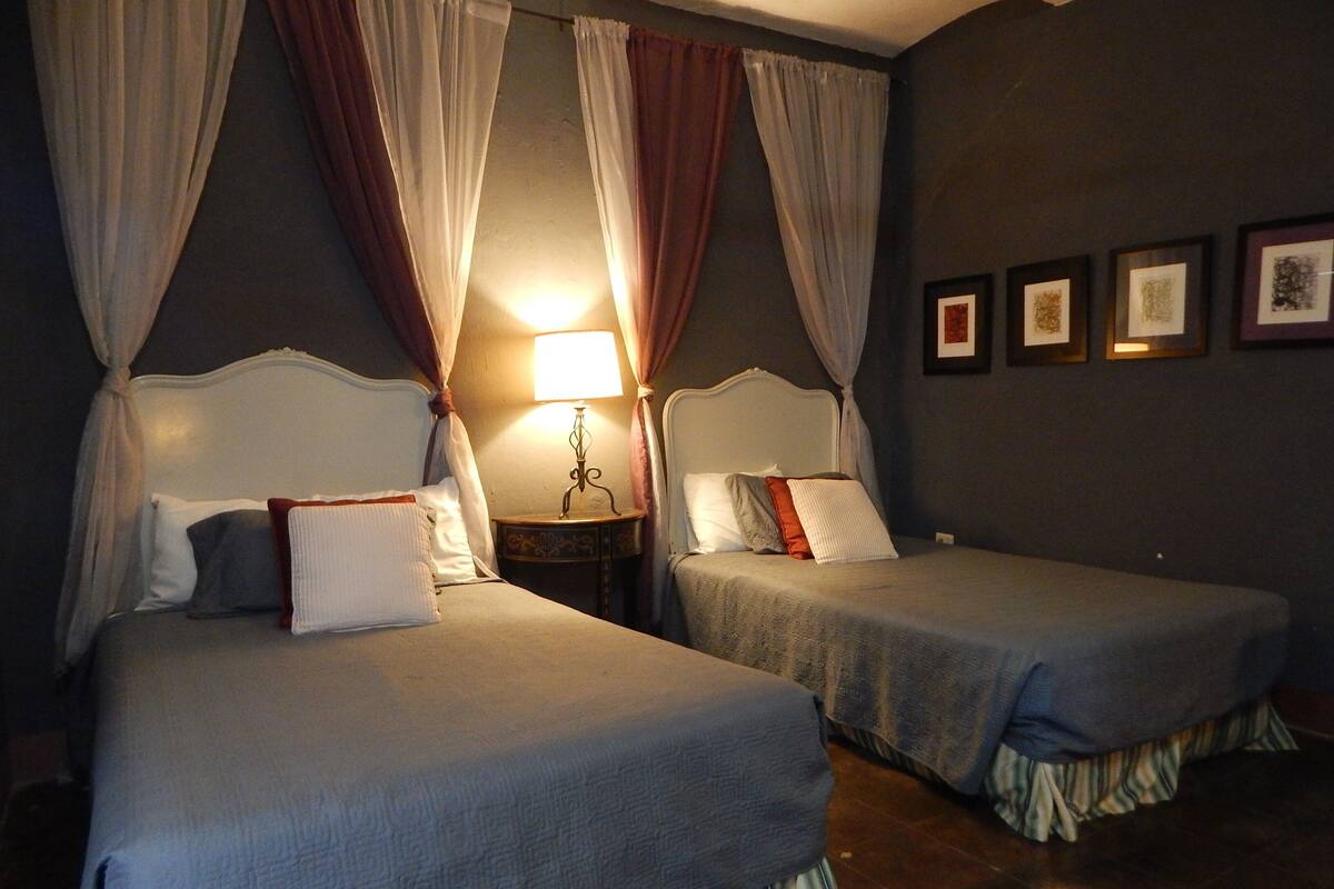 Een tweepersoonskamer met twee bedden, elk met hemelbed, nachtkastje met lamp en ingelijste foto’s aan de muur.