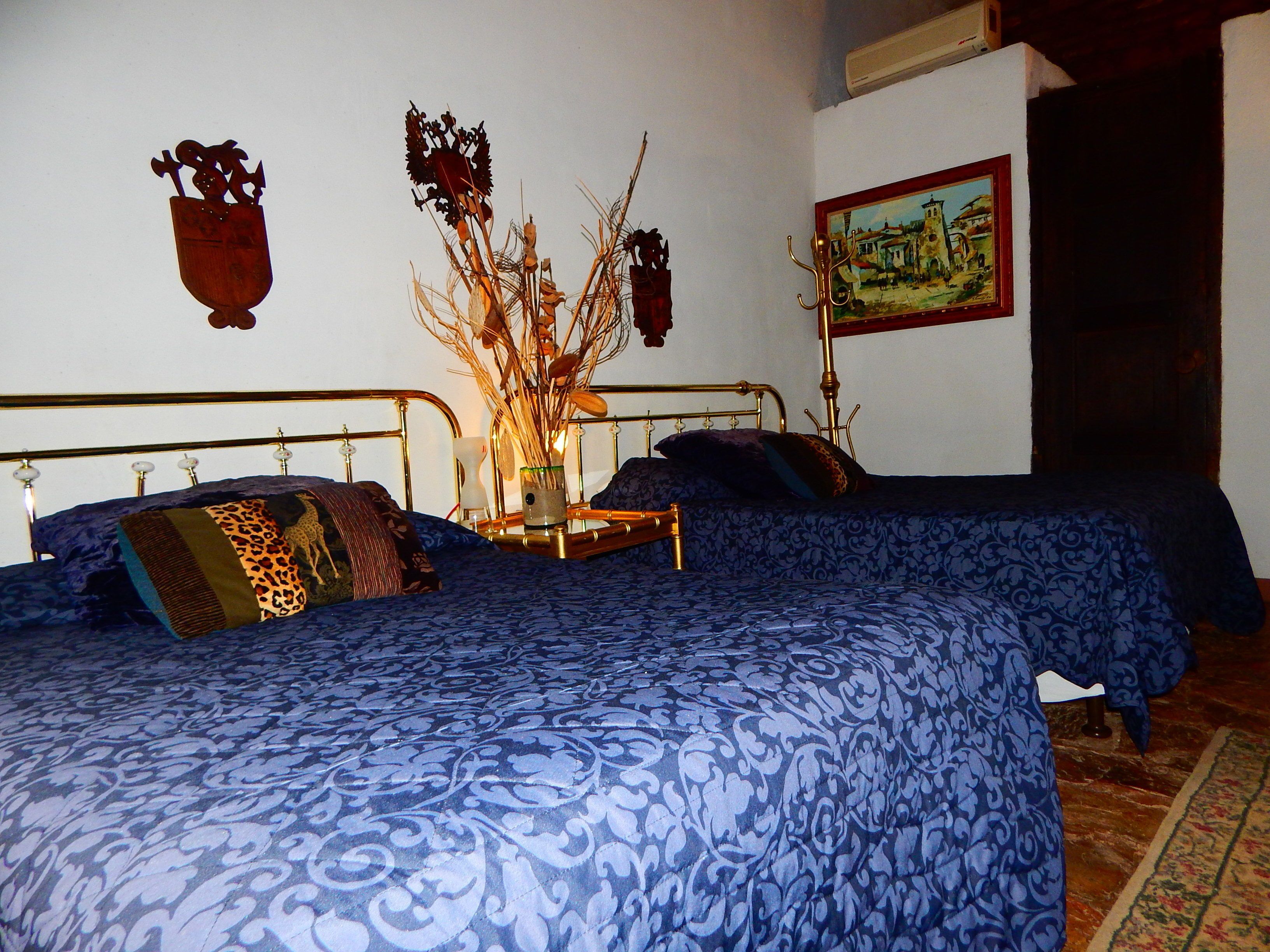 A room with two beds, each with a blue patterned bedspread and a wooden headboard. There is a wooden side table with a vase and a candle between the beds.