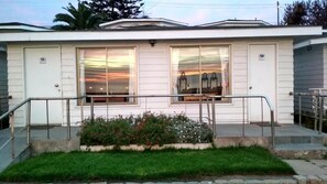 Un edificio de dos pisos con techo plano, dos ventanas grandes con cortinas, un pequeño porche con baranda de metal y un jardín con flores.