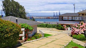 Un barrio residencial con un camino de entrada pavimentado, una casa pequeña con chimenea y vistas a un puerto con grúas al fondo.