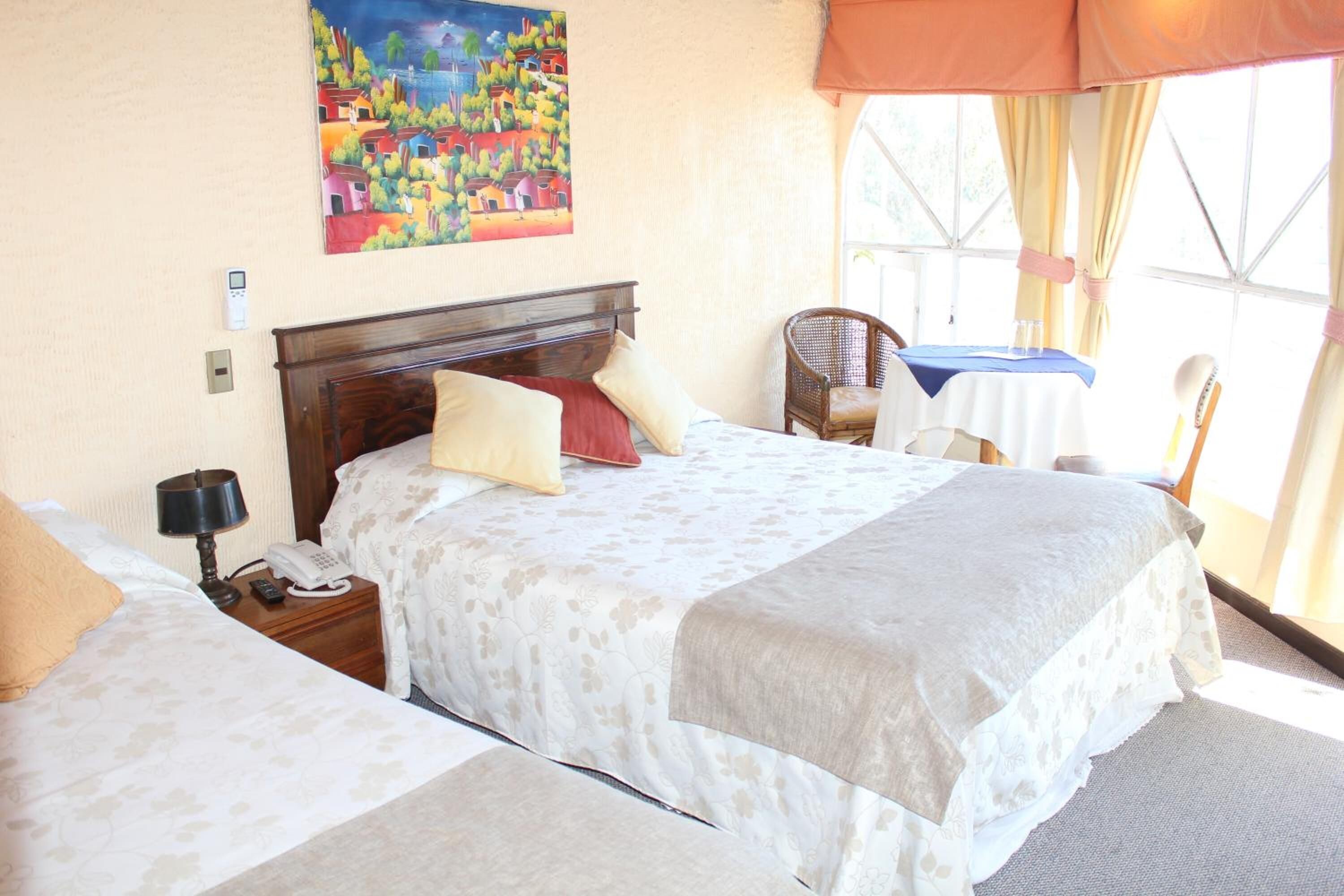 A hotel room with two beds, a painting on the wall, a window with curtains, and a small table with a chair.