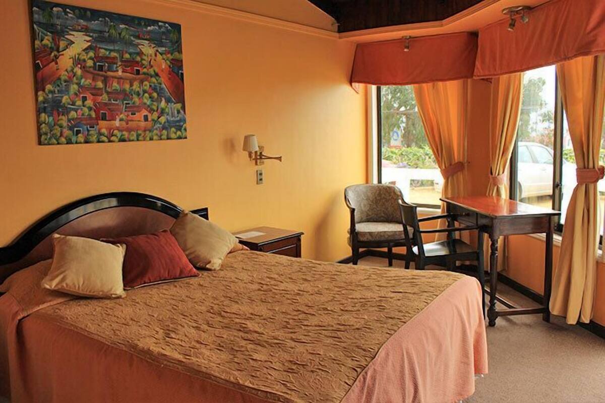 A hotel room with a bed, a desk, a chair, a painting on the wall, and a window with curtains.