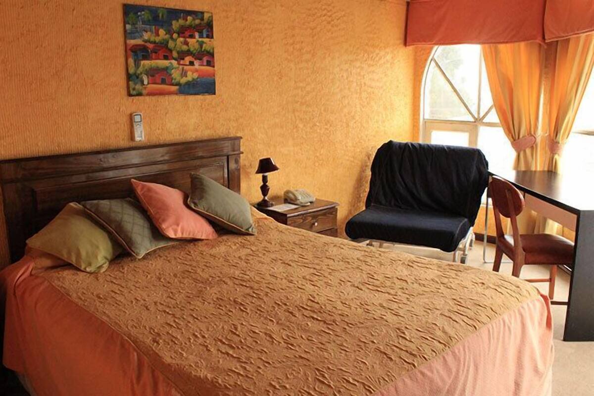 A bedroom with a bed, a desk, a chair, a nightstand, and a painting on the wall.