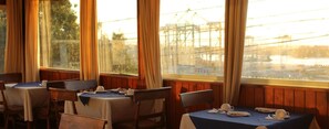Un restaurante con paredes de madera, mesas dispuestas con manteles blancos y vistas a un puente y al agua.