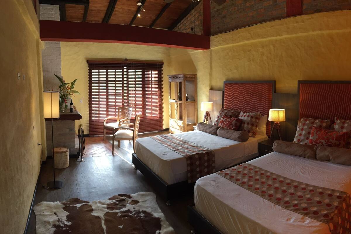 A hotel room with two beds, a large window with blinds, a wooden ceiling, and a small table with a lamp.
