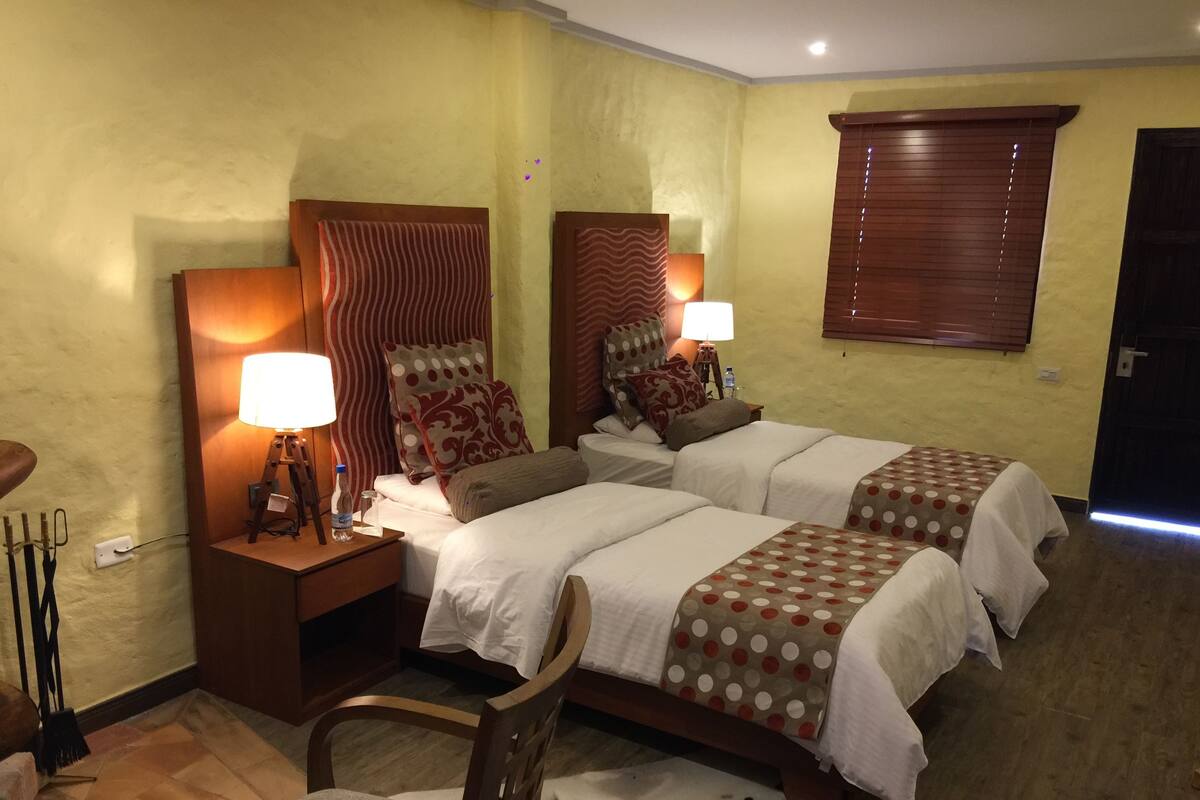 A hotel room with two beds, wooden headboards, and bedside lamps. There is a chair and a small rug with a cow print design.