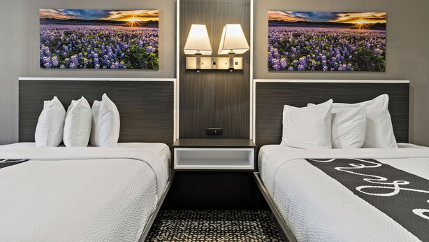 A hotel room with two beds, a nightstand with lamps, a mirror, and two framed pictures of a field at sunset.