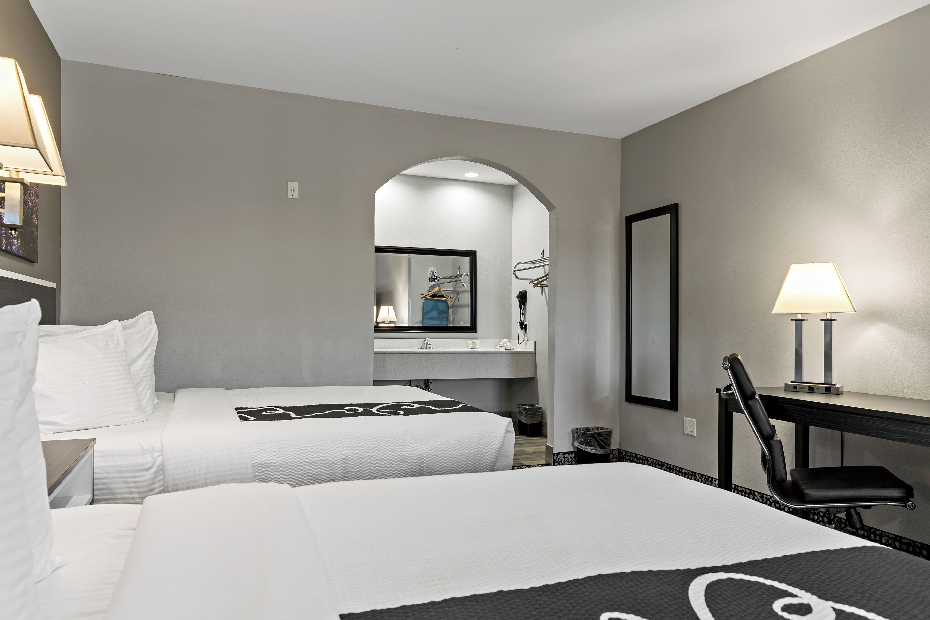 A hotel room with two beds, a desk, a chair, a mirror, and a lamp.