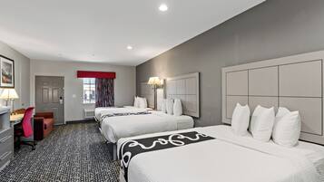 A hotel room with three beds, a desk with a chair, a window with curtains, and a lamp.