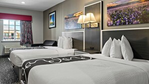 A hotel room with two beds, a large window with curtains, a painting of flowers, and a wall-mounted lamp.
