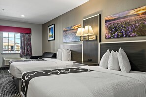 A hotel room with two beds, a large window with curtains, a painting of flowers, and a wall-mounted lamp.