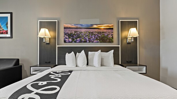 A hotel room with a large bed, two bedside tables, a headboard with a sunset painting, and two wall-mounted lamps.