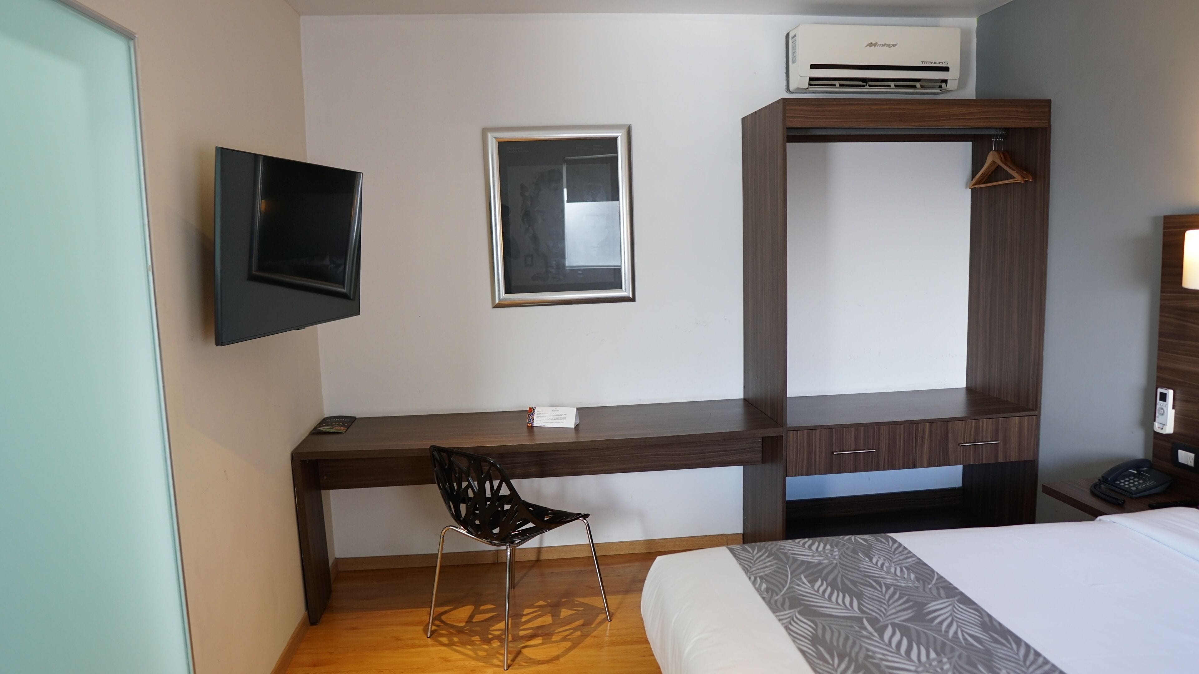 A hotel room with a wooden desk, a chair, a bed, a TV mounted on the wall, and a wardrobe.
