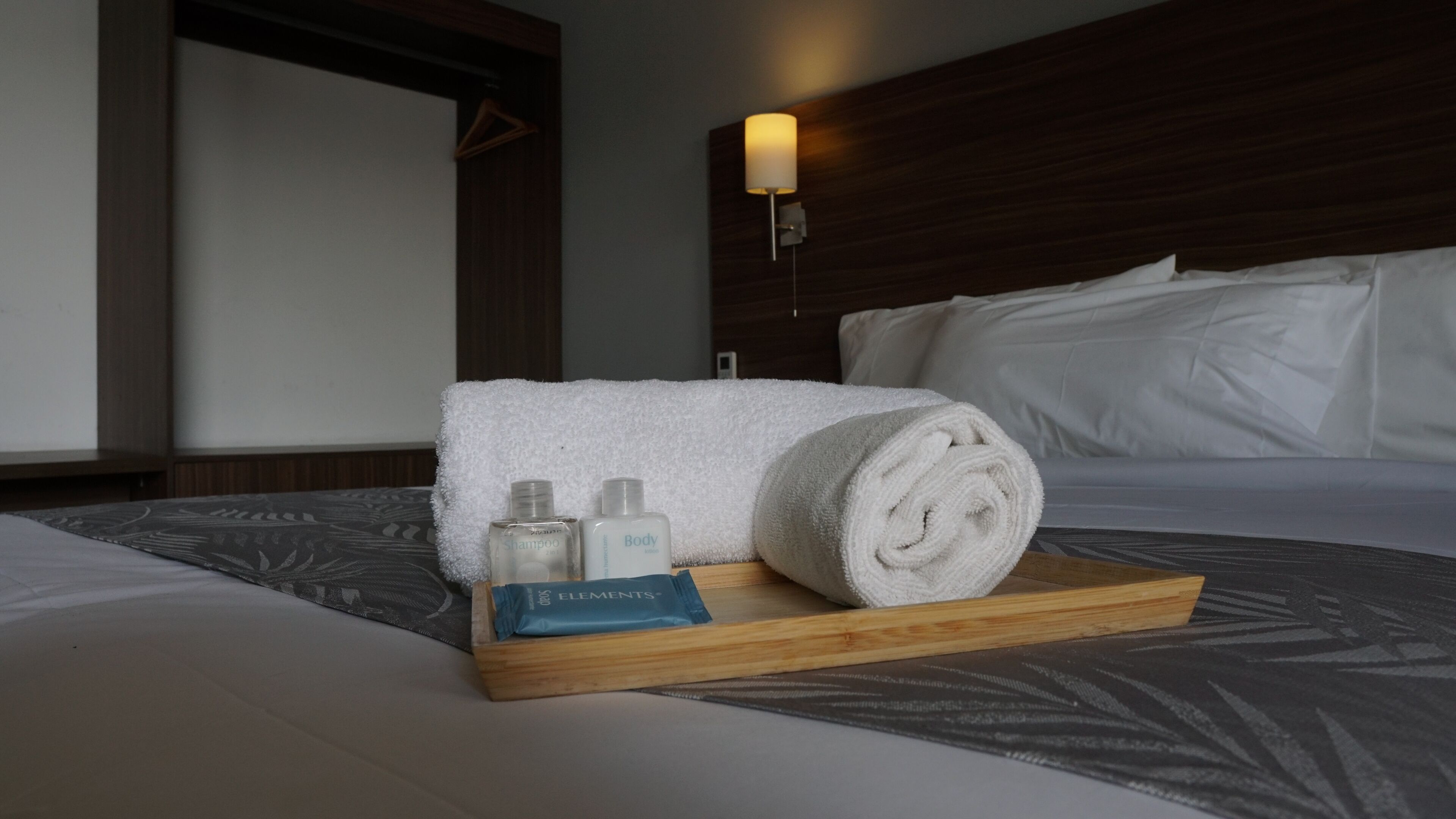 A neatly made bed with a wooden tray holding a rolled towel, two bottles of shampoo, and a bar of soap.