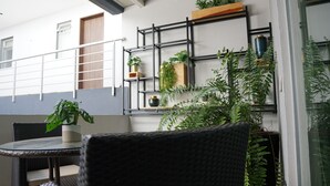 Un área de estar moderna al aire libre con una mesa redonda y sillas de mimbre, rodeada de plantas en macetas y una estantería metálica.