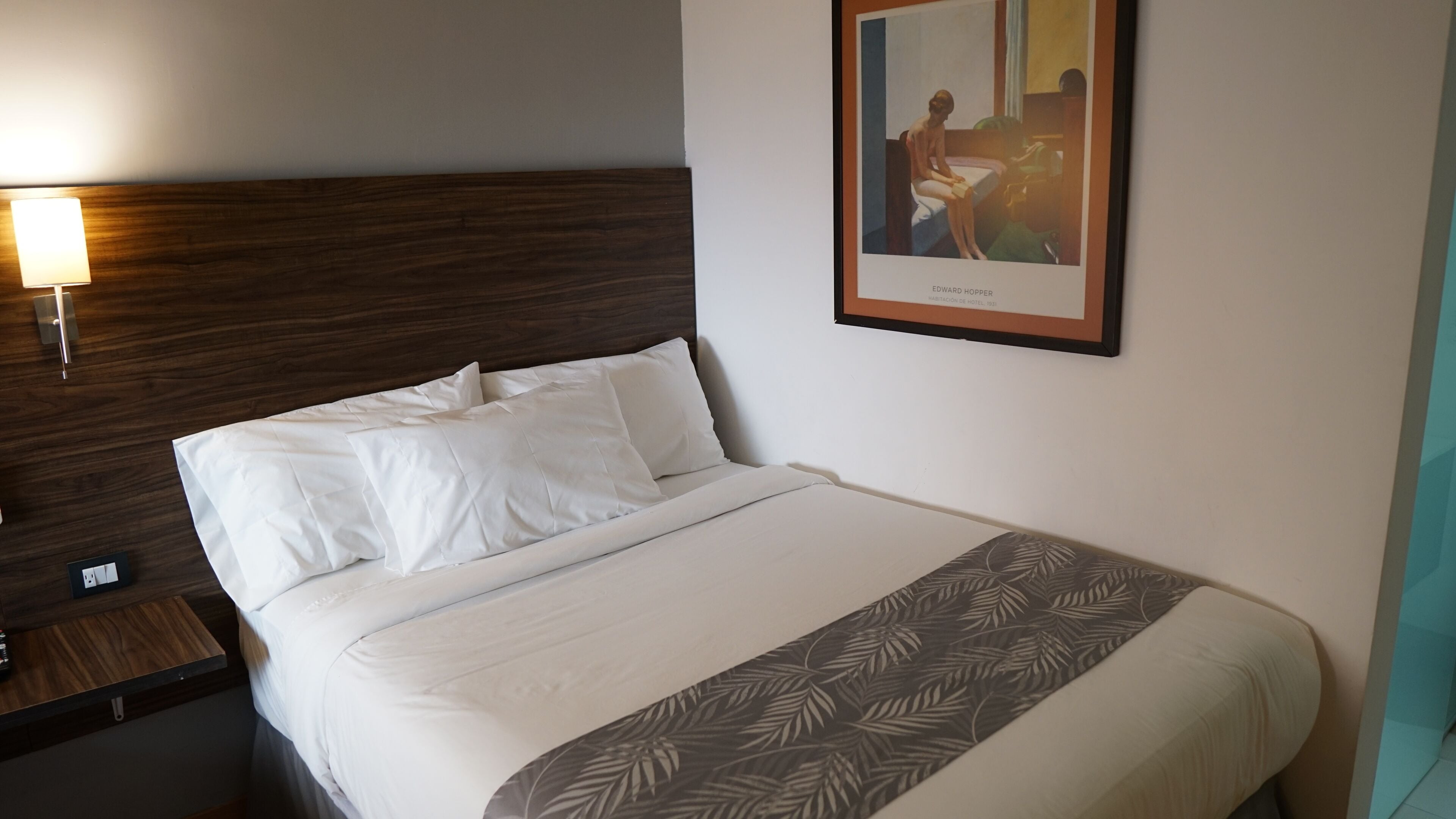 A hotel room with a neatly made bed, a nightstand with a lamp, and a framed painting on the wall.