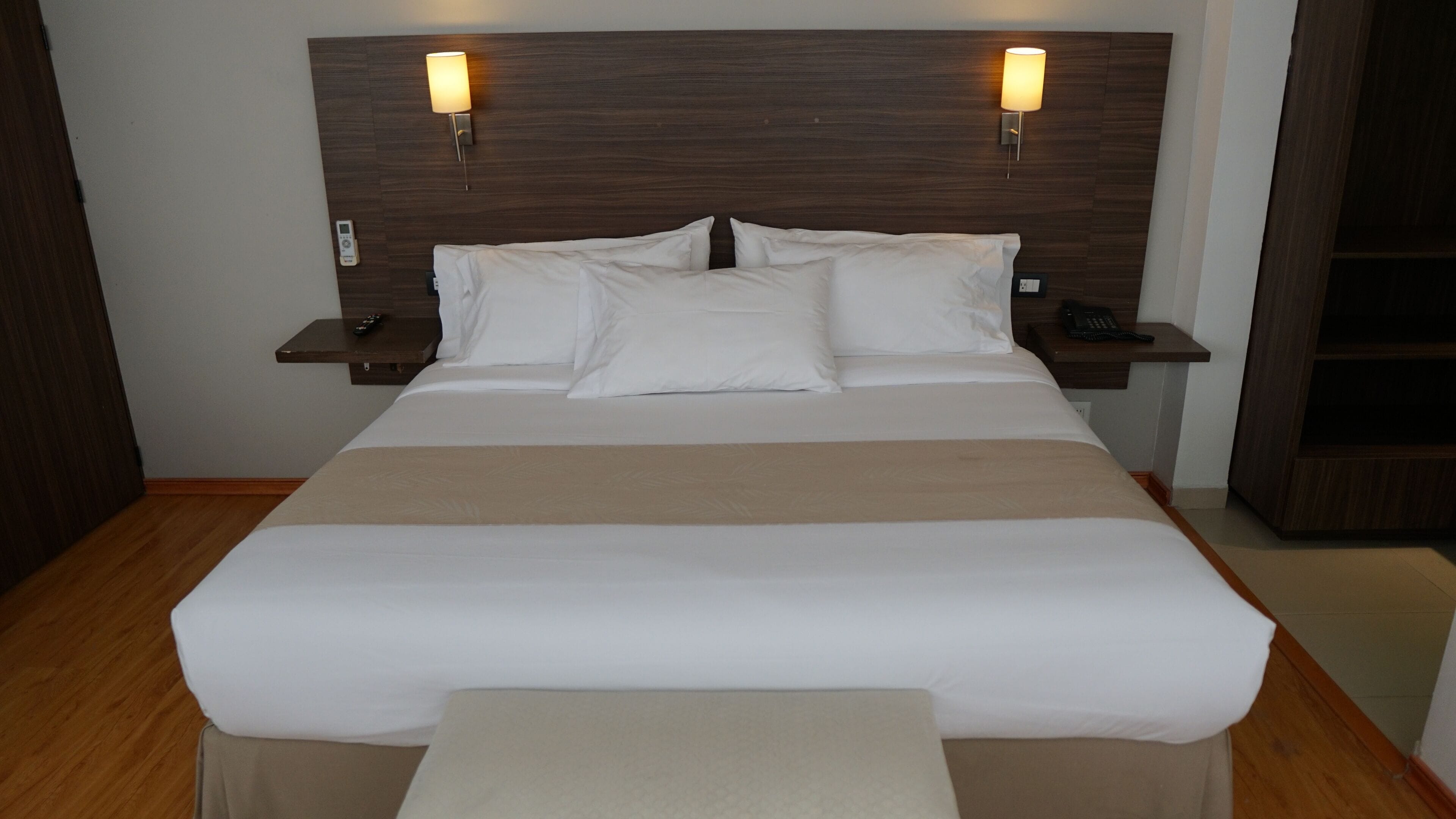 Una cama bien hecha con sábanas blancas y un plaid color beige, flanqueada por dos mesitas de noche con lámparas, un control remoto y un teléfono.
