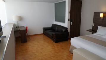 Una habitación de hotel con una cama, un escritorio con lámpara, un sofá de cuero y suelo de madera.