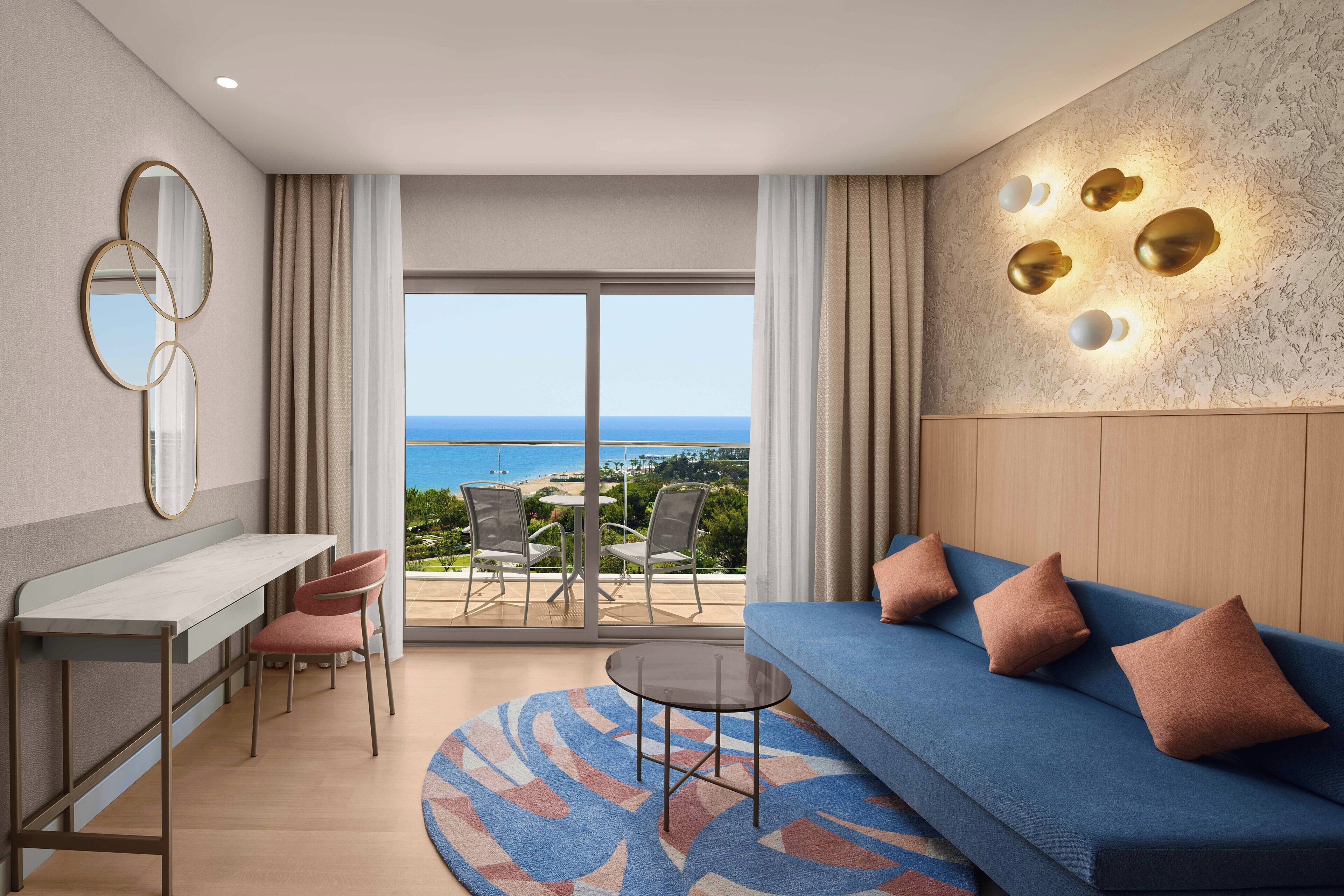 A modern living room with a sofa, a small table, and a desk. There is a view of the ocean through large windows.