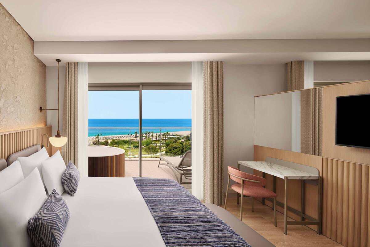 A hotel room with a large bed, a desk, a chair, and a TV. There is a balcony with a view of the beach and ocean.