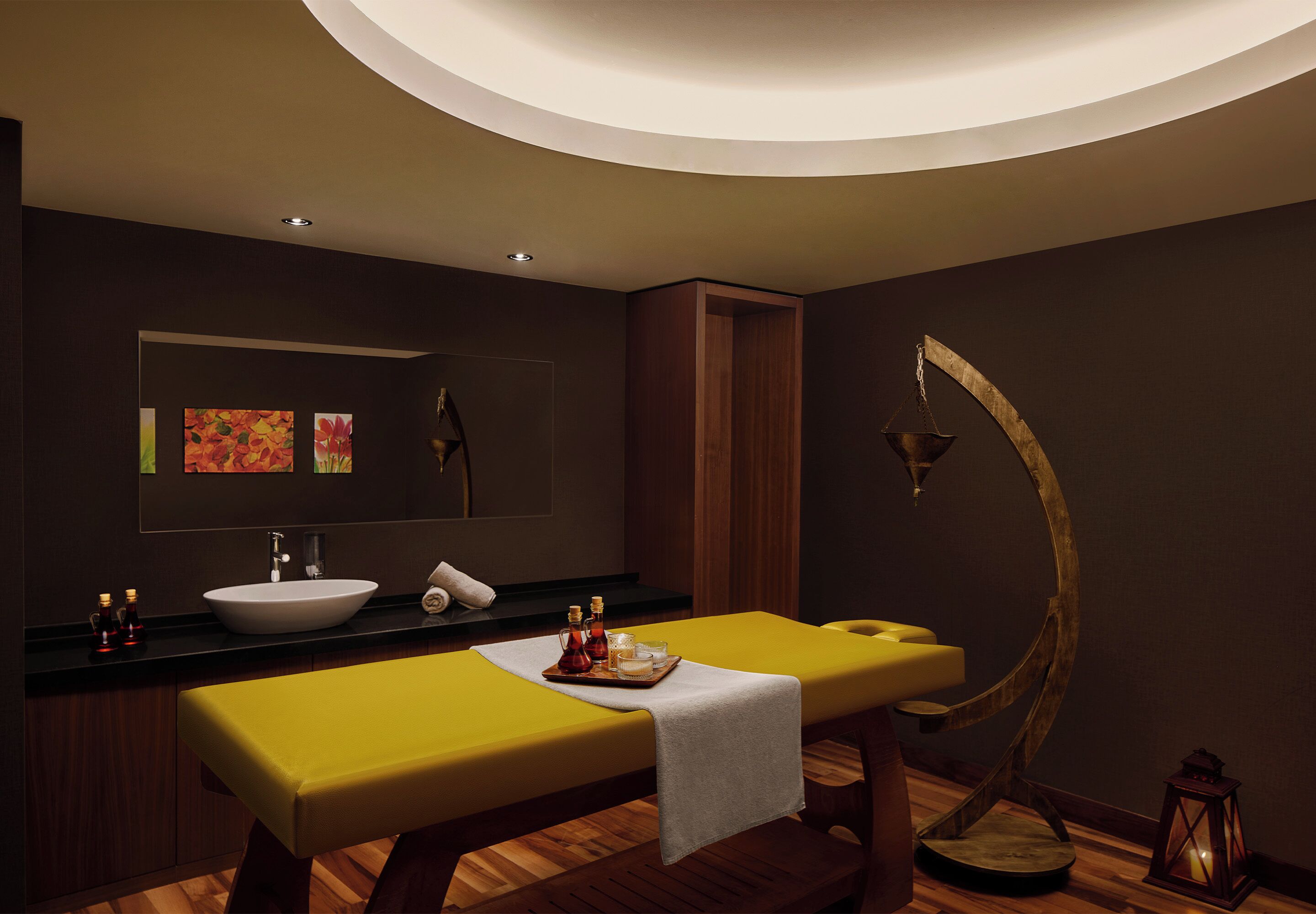 A modern spa room with a wooden massage table, a large mirror, a sink, and a wooden arch light fixture.