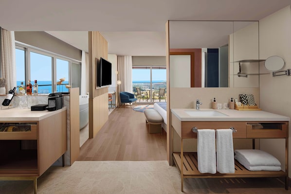 Una habitación de hotel moderna con un ventanal grande, un balcón con vista al mar, un televisor de pantalla plana, un escritorio de madera y un baño con un espejo grande y un perchero para toallas.