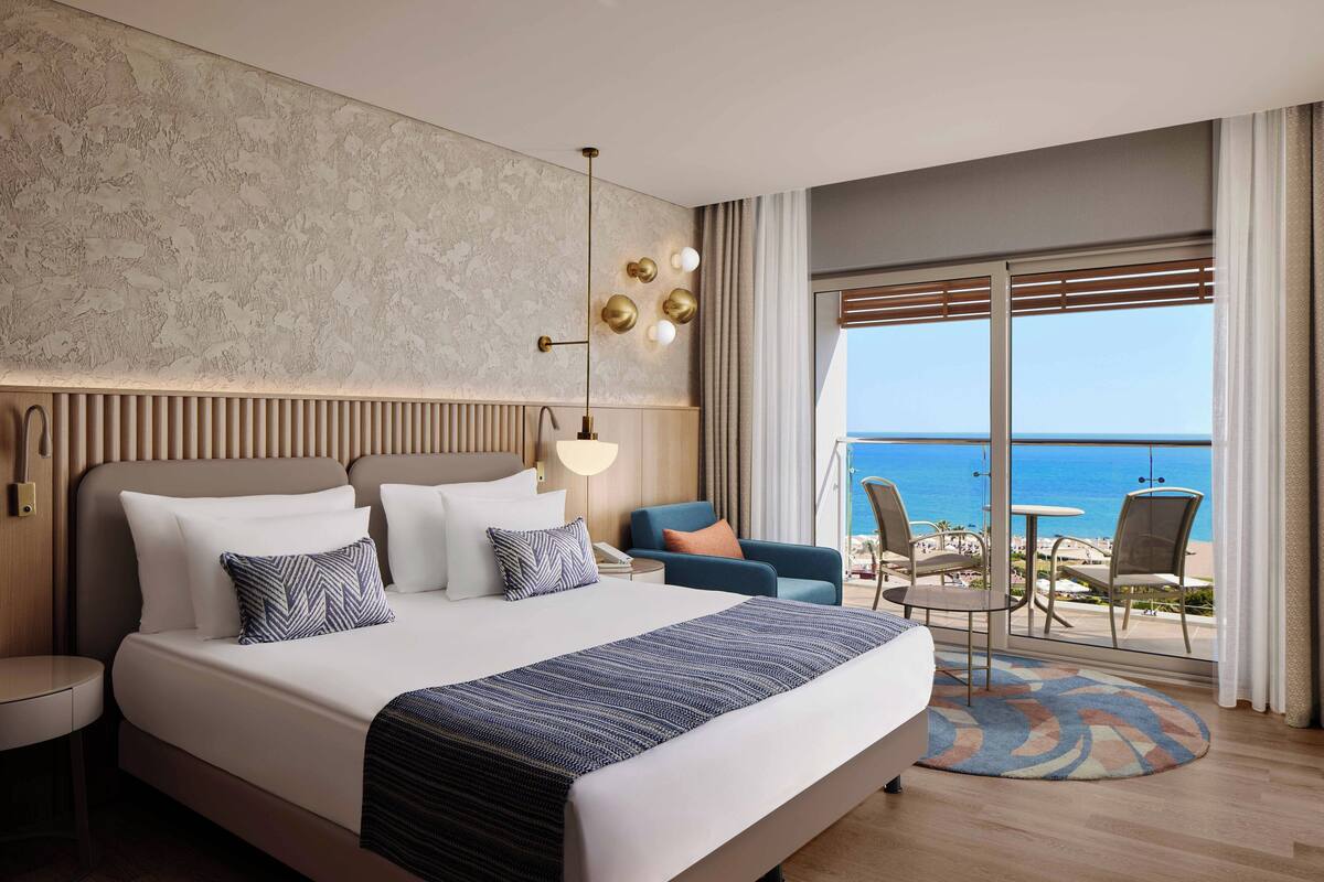 A modern hotel room with a large bed, a view of the sea, and a balcony with seating.
