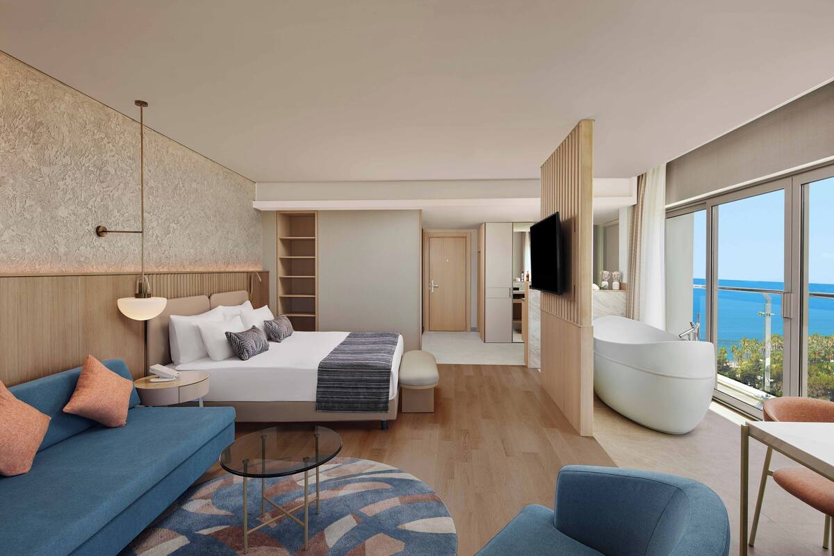 A modern hotel room with a large bed, a sofa, a flat-screen TV, a bathtub, and a balcony with a view of the sea.