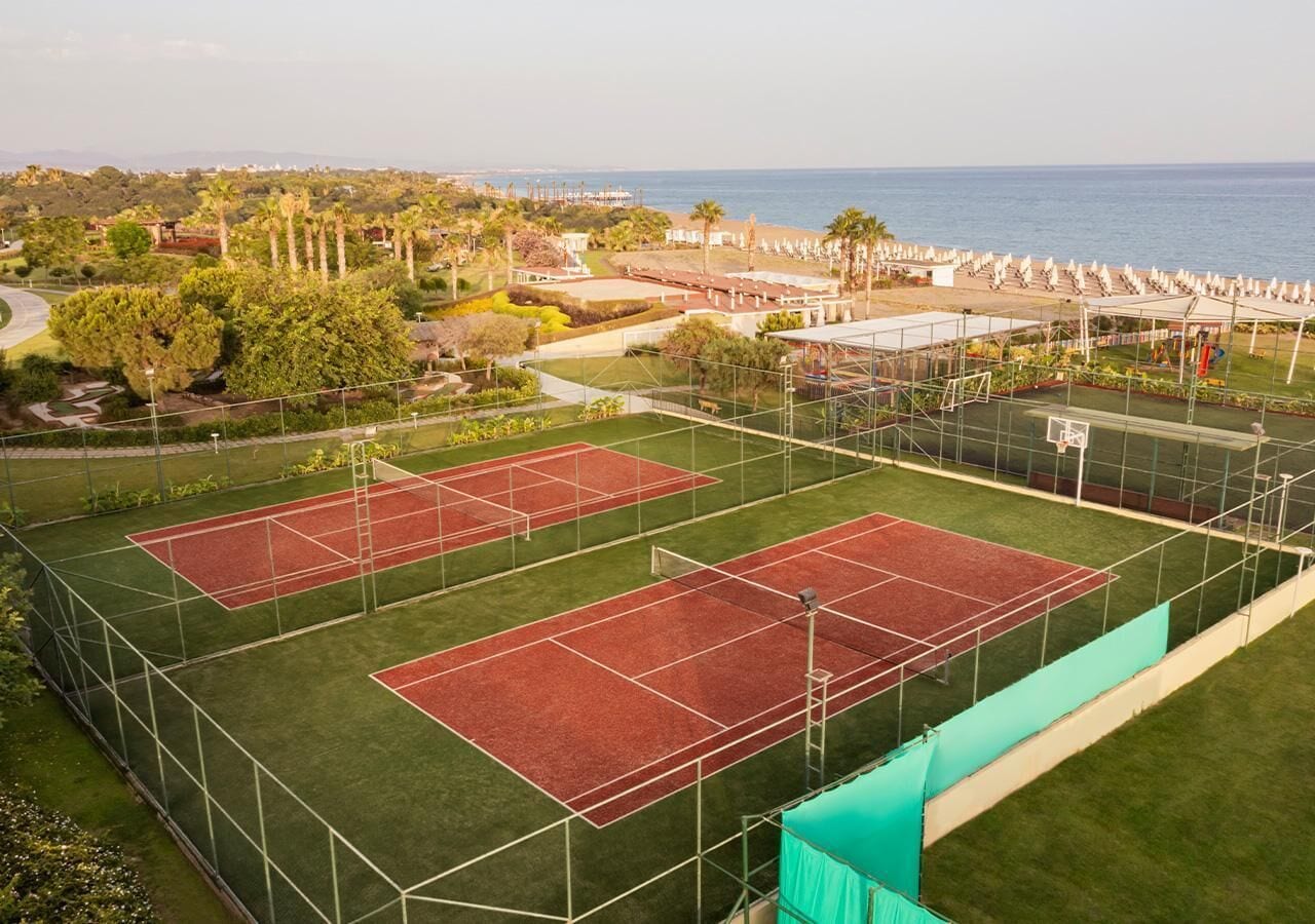 An outdoor tennis court complex with multiple courts, surrounded by greenery and a view of the ocean.