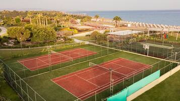 An outdoor tennis court complex with multiple courts, surrounded by greenery and a view of the ocean.