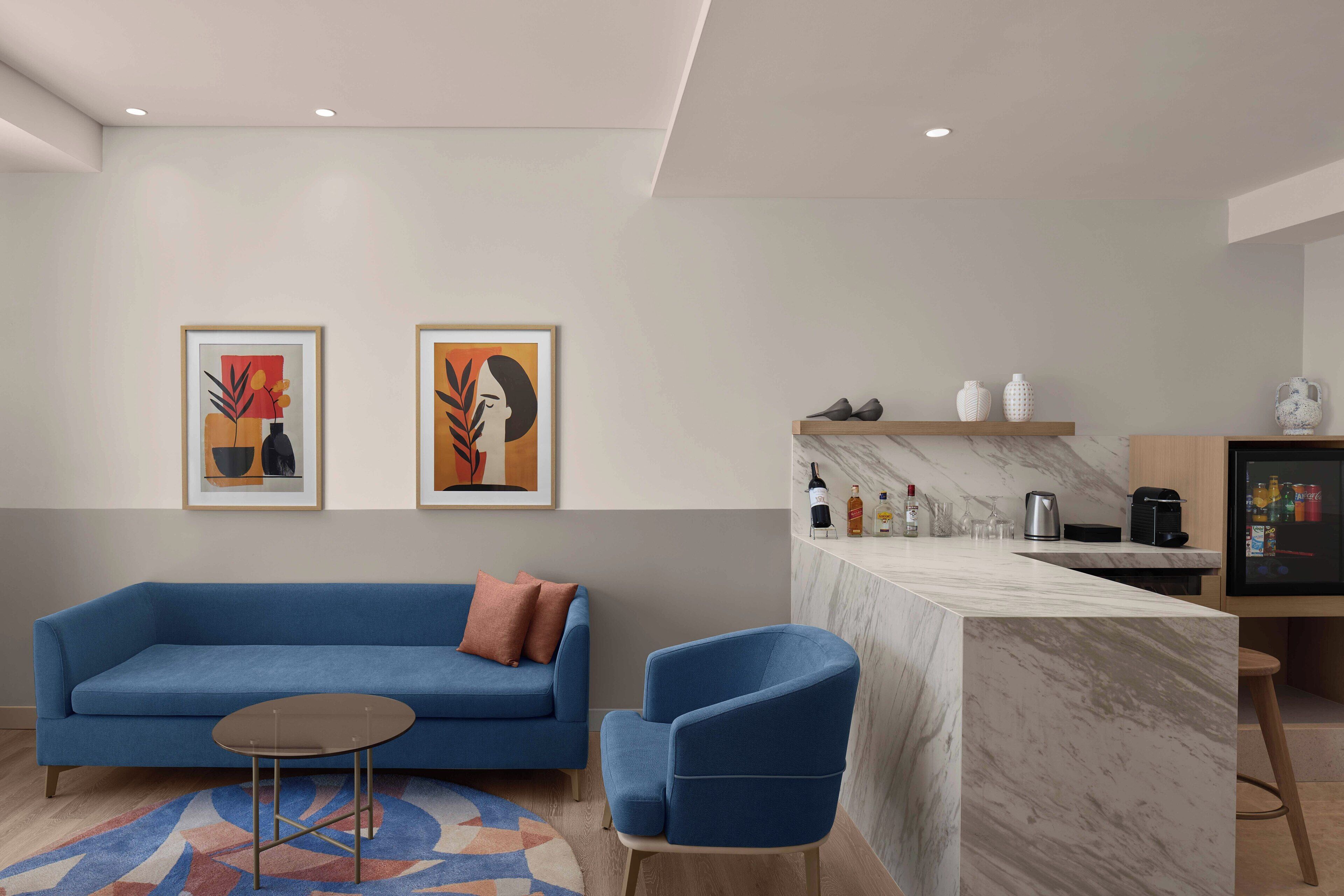 Un salón moderno con un sofá azul, una pequeña mesa redonda y un sillón azul. Hay dos obras de arte enmarcadas en la pared, una zona de cocina con encimera de mármol y un refrigerador lleno de bebidas.