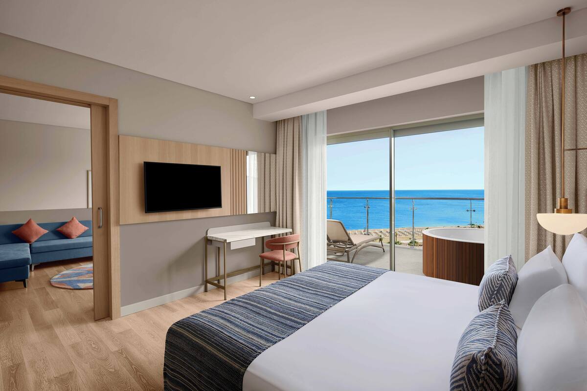 A hotel room with a bed, desk, TV, and a balcony with a view of the ocean.