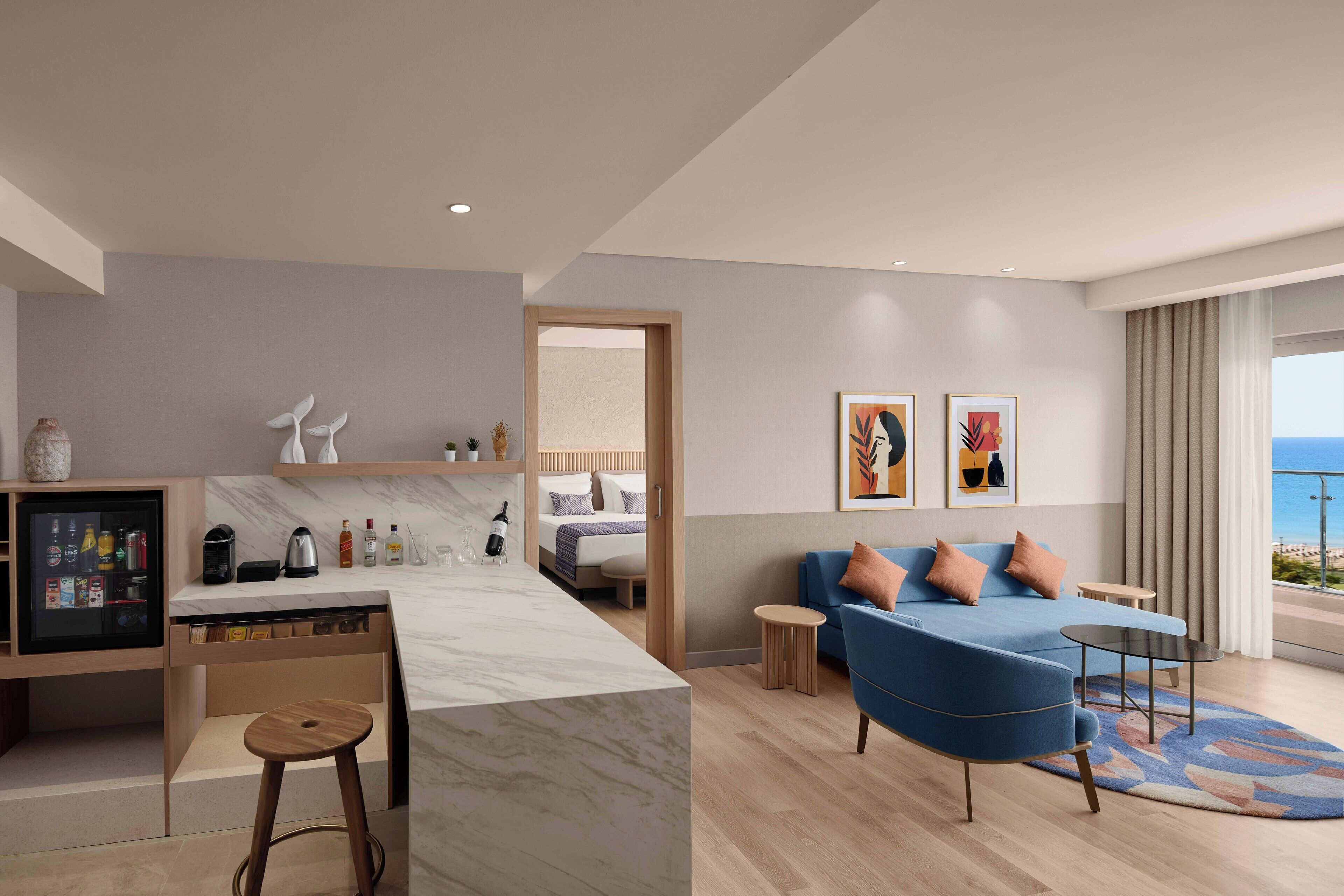 A modern hotel room with a kitchen area, a sofa set, and a balcony with a view.