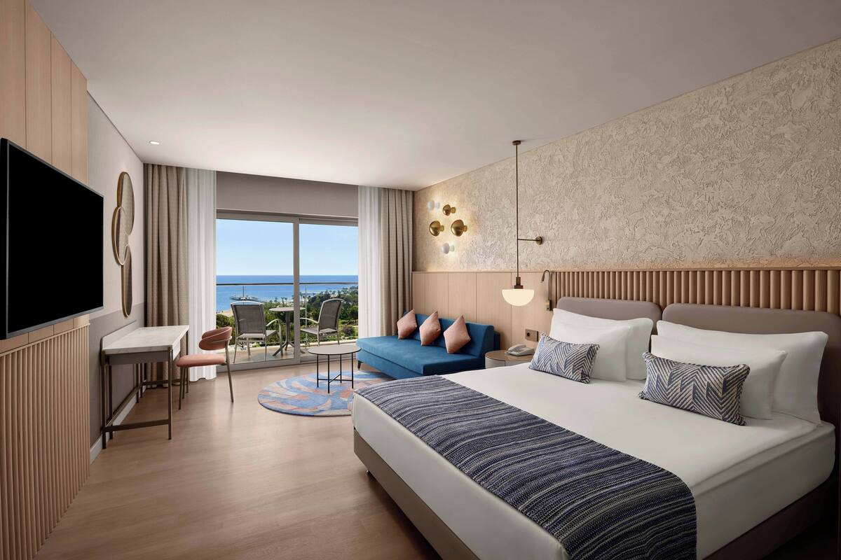 A modern hotel room with a large bed, a desk, a chair, and a balcony with a view of the sea.